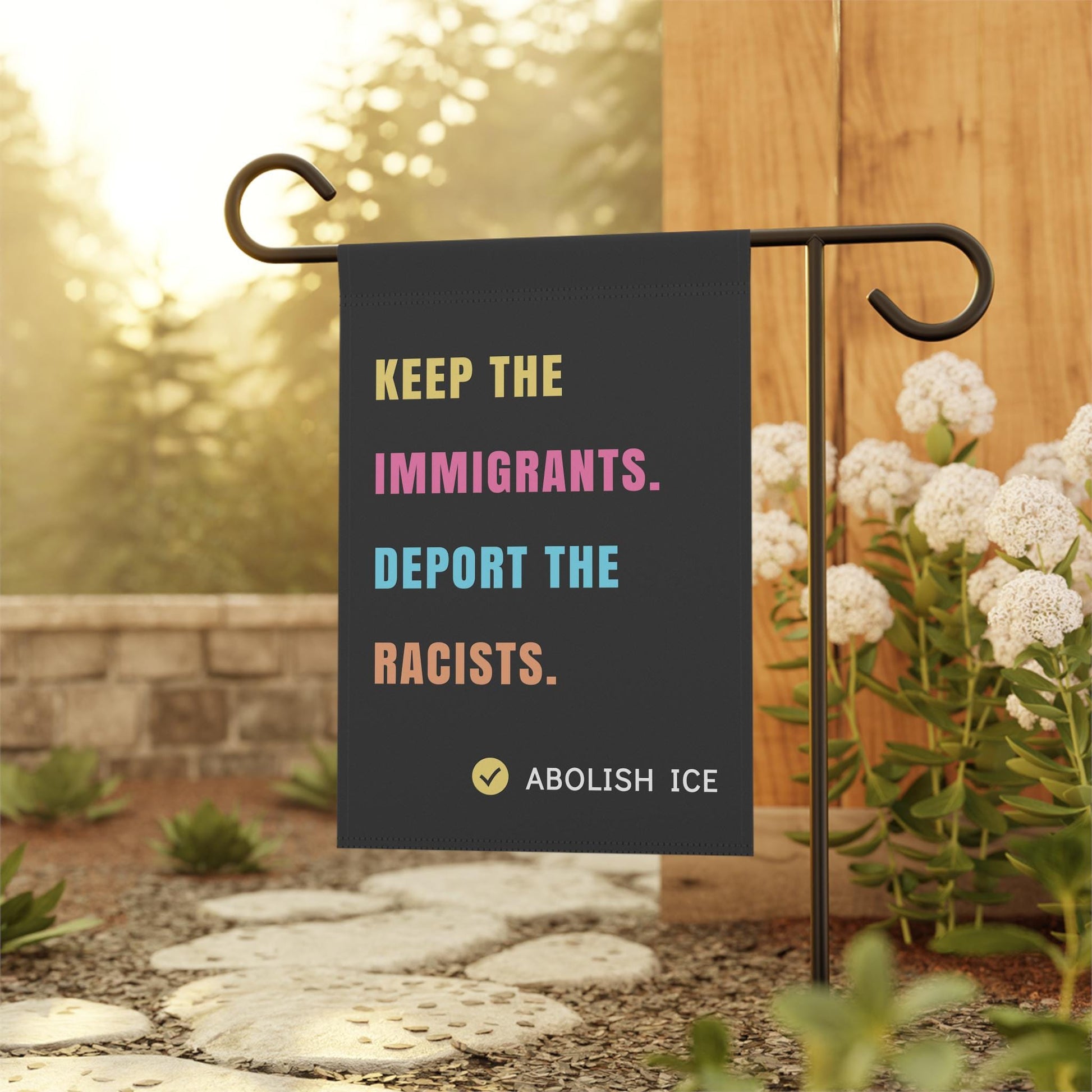 Black garden flag with bold, multicolored text reading "KEEP THE IMMIGRANTS. DEPORT THE RACISTS. ABOLISH ICE," displayed outdoors on a metal flag stand surrounded by flowers and greenery. Pro-immigrant, anti-racist activist flag for yard or garden decor.