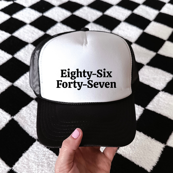 A hand holding a black and white trucker hat with the text 'Eighty-Six Forty-Seven' printed on the front, set against a black and white checkered rug.