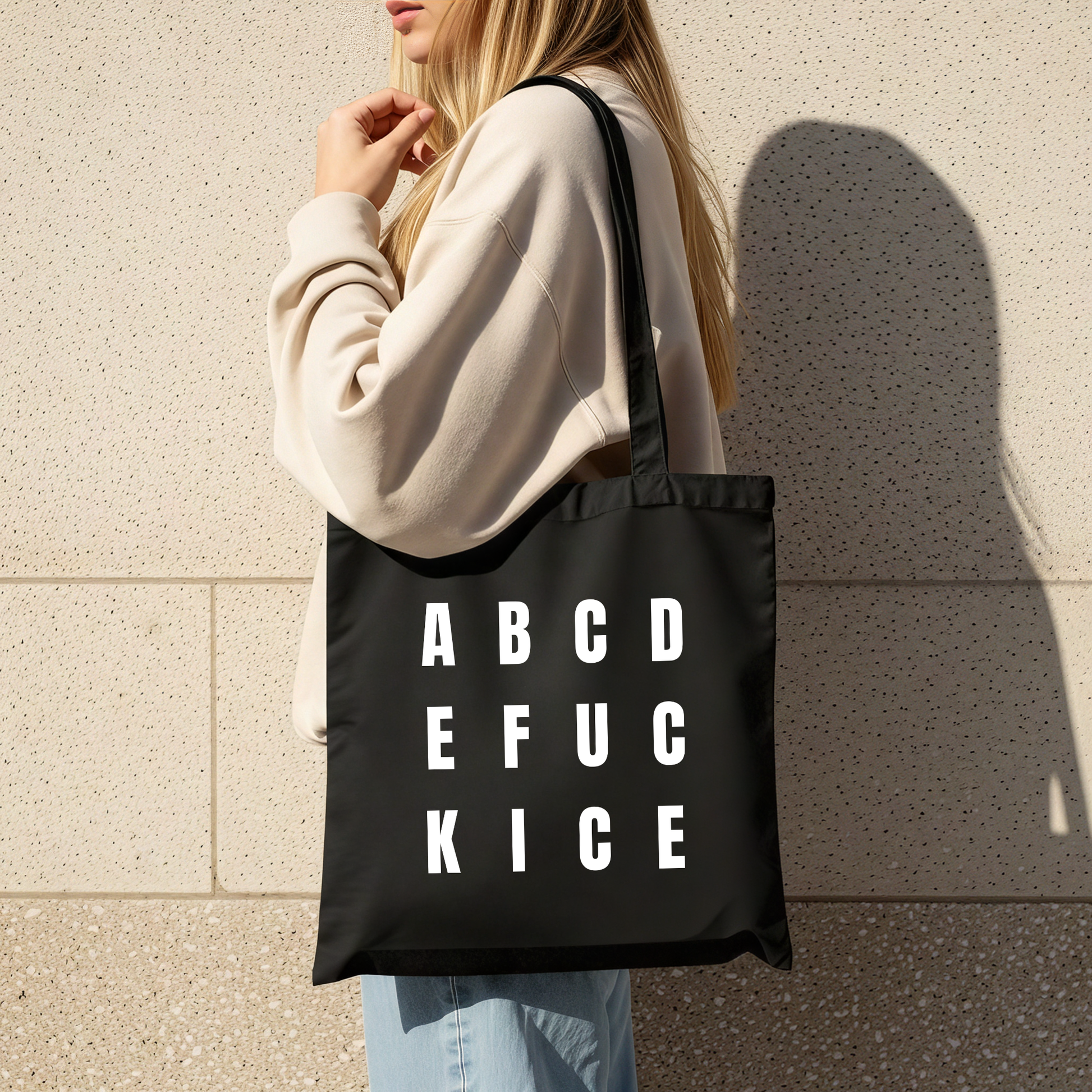 Person holding a black tote bag with white text reading ABCDE FUCK ICE against a beige wall.