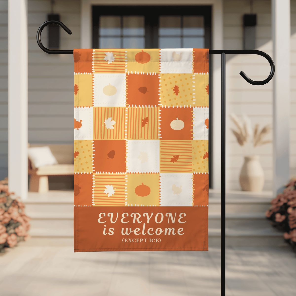 An autumn-themed garden flag hanging from a black metal stand on a front porch. The flag has a patchwork design in orange, yellow, and white with fall motifs like pumpkins, leaves, and stripes, and a bottom banner that reads 'EVERYONE is welcome (EXCEPT ICE)'.
