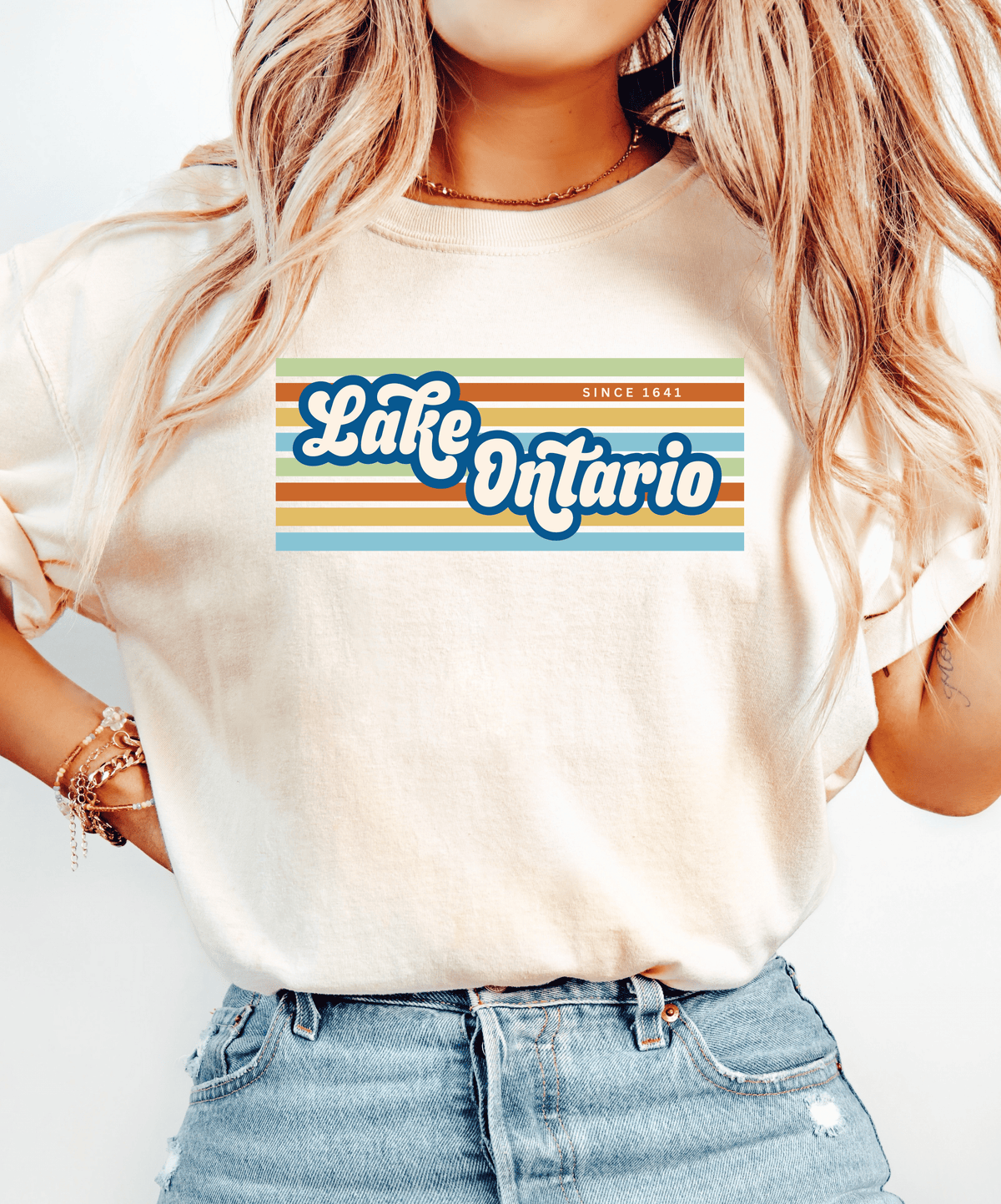 A woman wearing a cream-colored t-shirt with a retro-style graphic that reads 'Lake Ontario Since 1641' in blue and white lettering over horizontal orange, yellow, and green stripes. She is also wearing blue jeans and a gold necklace.
