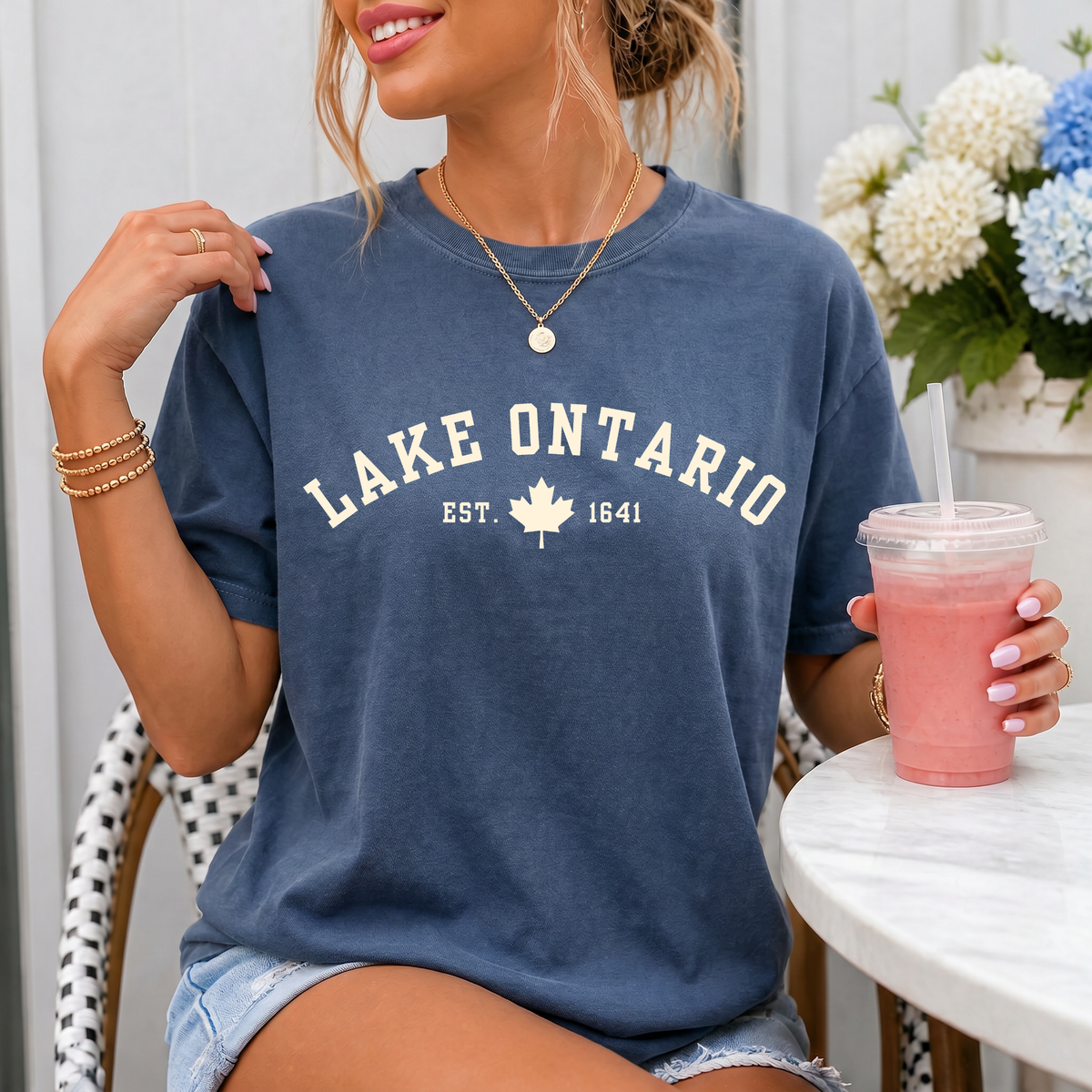 A woman wearing a faded blue crewneck t-shirt with 'LAKE ONTARIO EST. 1641' printed in white on the front, holding a pink drink next to a white table.