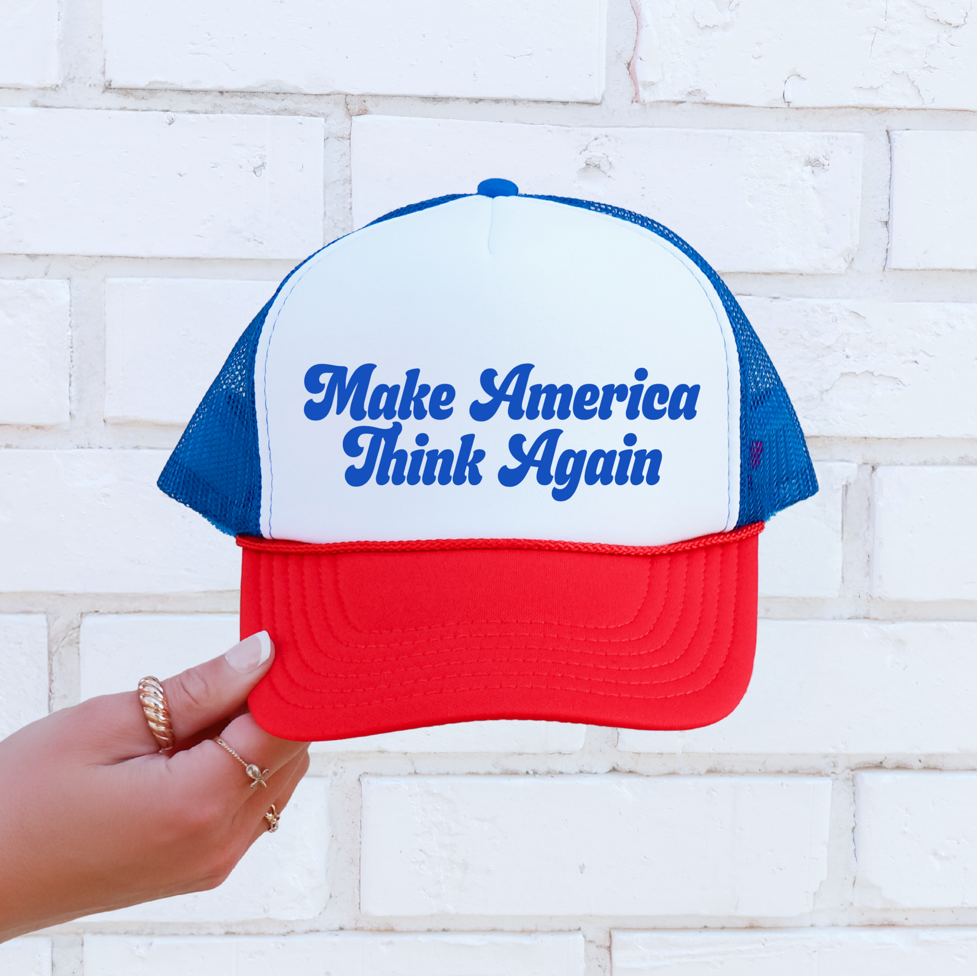 A red, white, and blue trucker hat with the text 'Make America Think Again' printed in blue on the front, held by a hand against a white brick wall background.