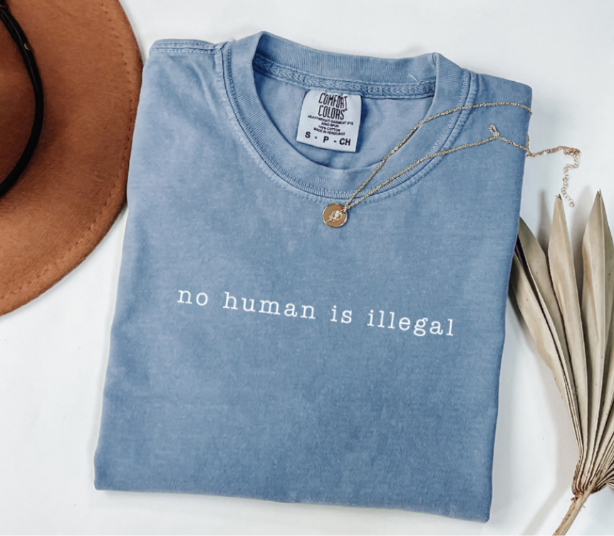 Folded blue t-shirt with 'no human is illegal' text on a white surface with a hat and dried leaves.