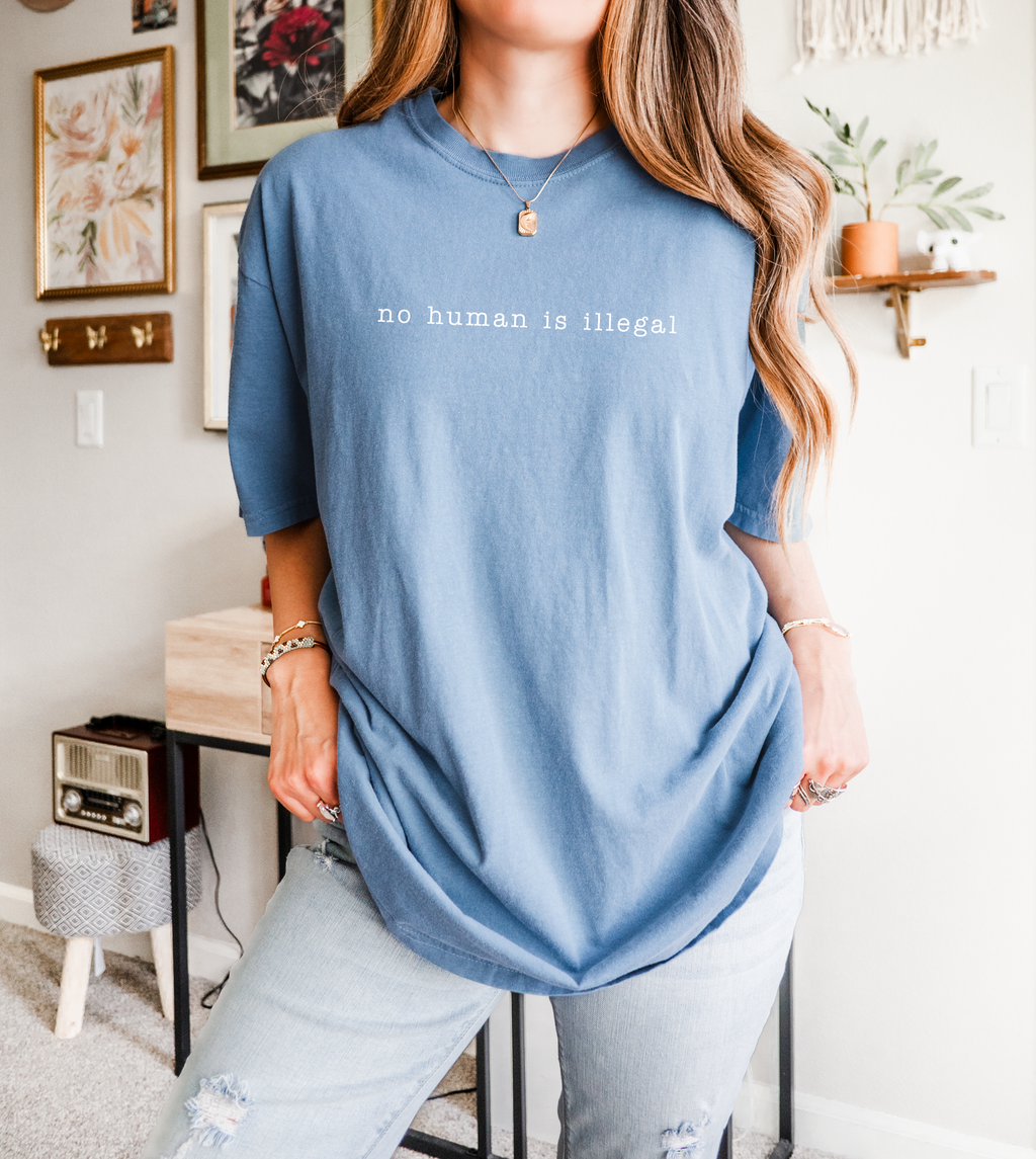 Person wearing a blue t-shirt with 'no human is illegal' text in a home setting.