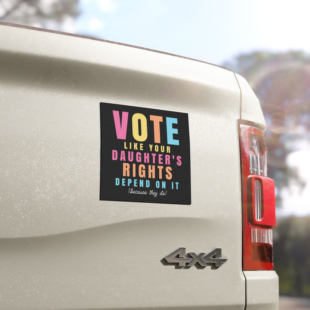 A rectangular black bumper sticker with colorful text reading 'VOTE LIKE YOUR DAUGHTER'S RIGHTS DEPEND ON IT (Because They Go)' applied to the rear side of a light-colored vehicle.
