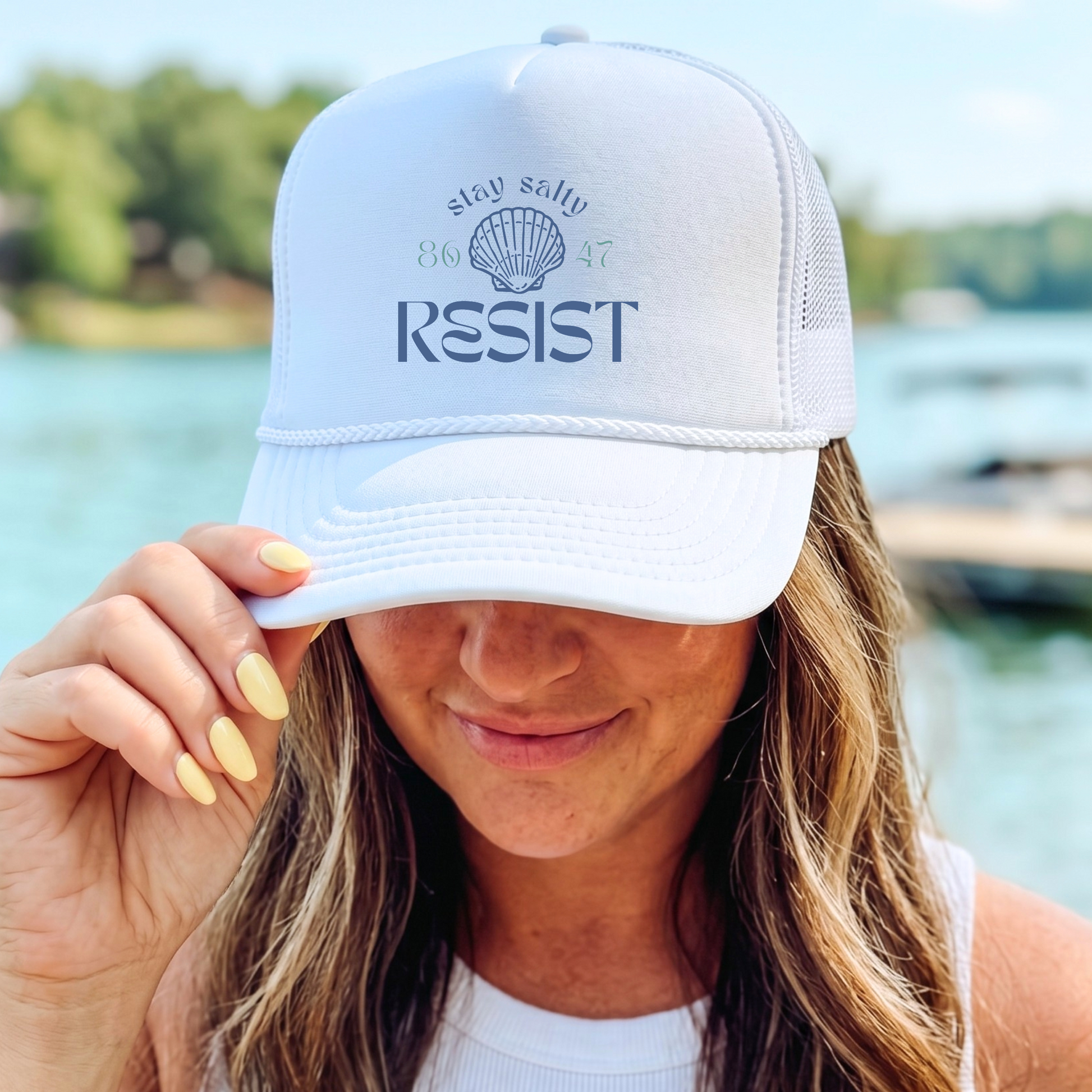 A woman wearing a white trucker hat with 'stay salty RESIST' and a seashell graphic printed on the front, holding the brim over her eye in front of a sunny lake background.