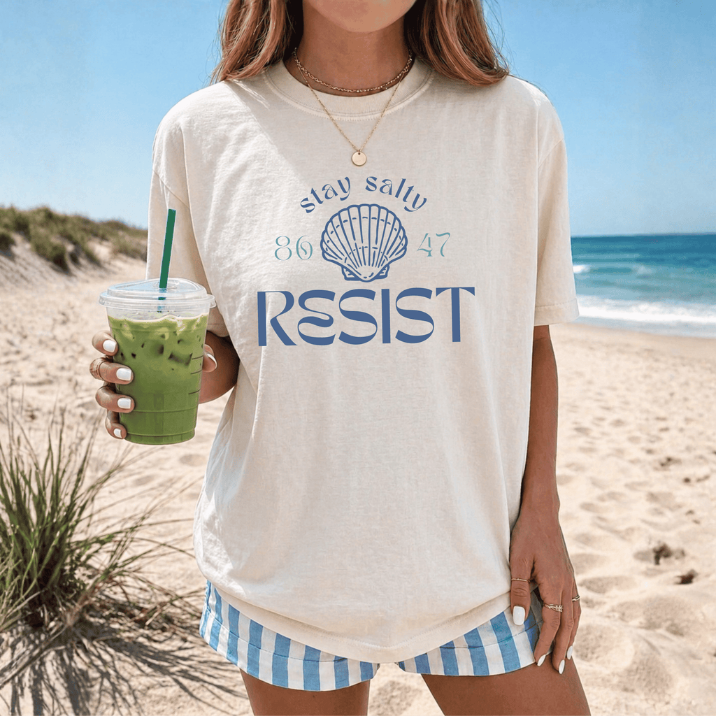 Person wearing a t-shirt with 'Stay salty 86 47 RESIST' design on a beach.