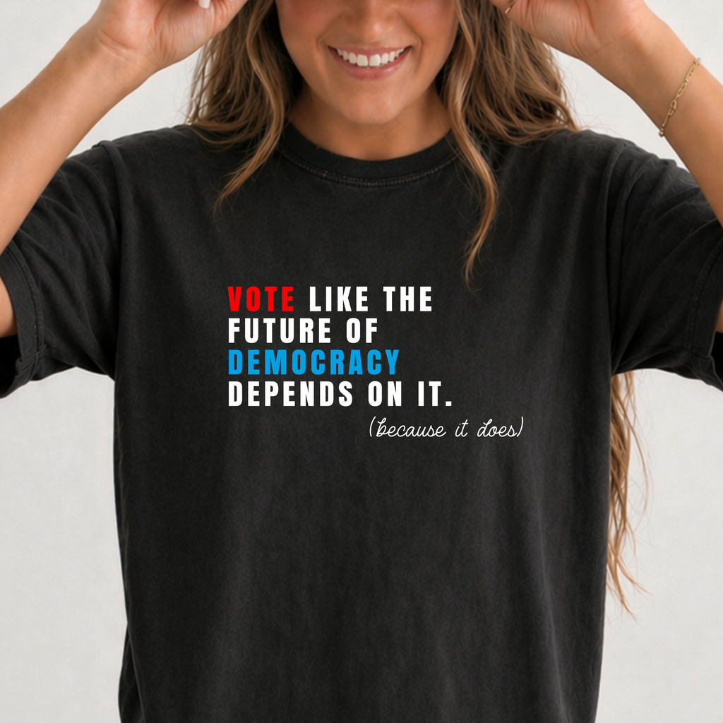 A woman sitting cross-legged wearing a black graphic t-shirt with a political slogan, blue jeans, and a beige baseball cap. The t-shirt features a quote in red, white, and blue text.