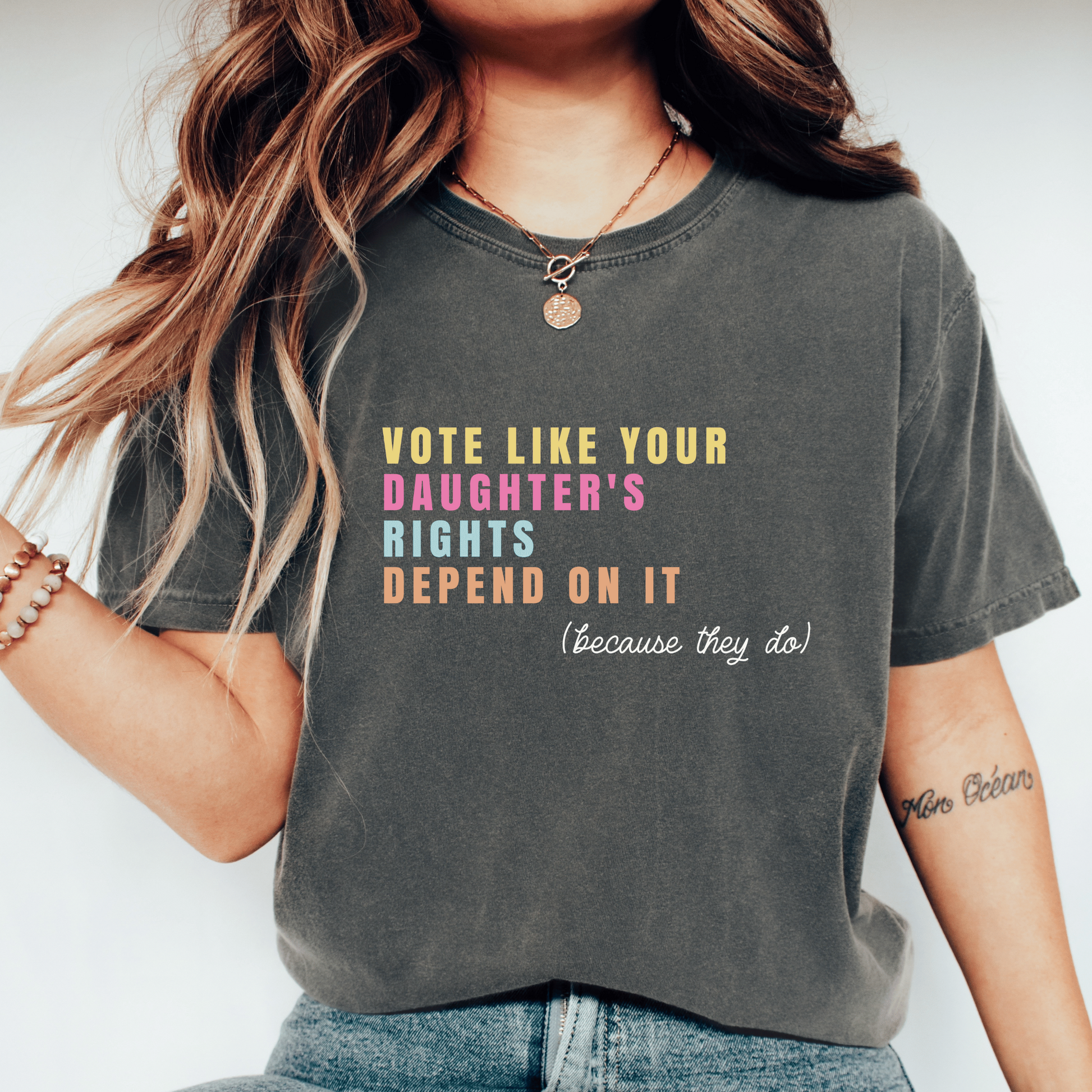 A woman wearing a dark gray graphic t-shirt with the text 'VOTE LIKE YOUR DAUGHTER'S RIGHTS DEPEND ON IT (because they do!)' in yellow, pink, and orange lettering, paired with blue jeans and white sneakers.